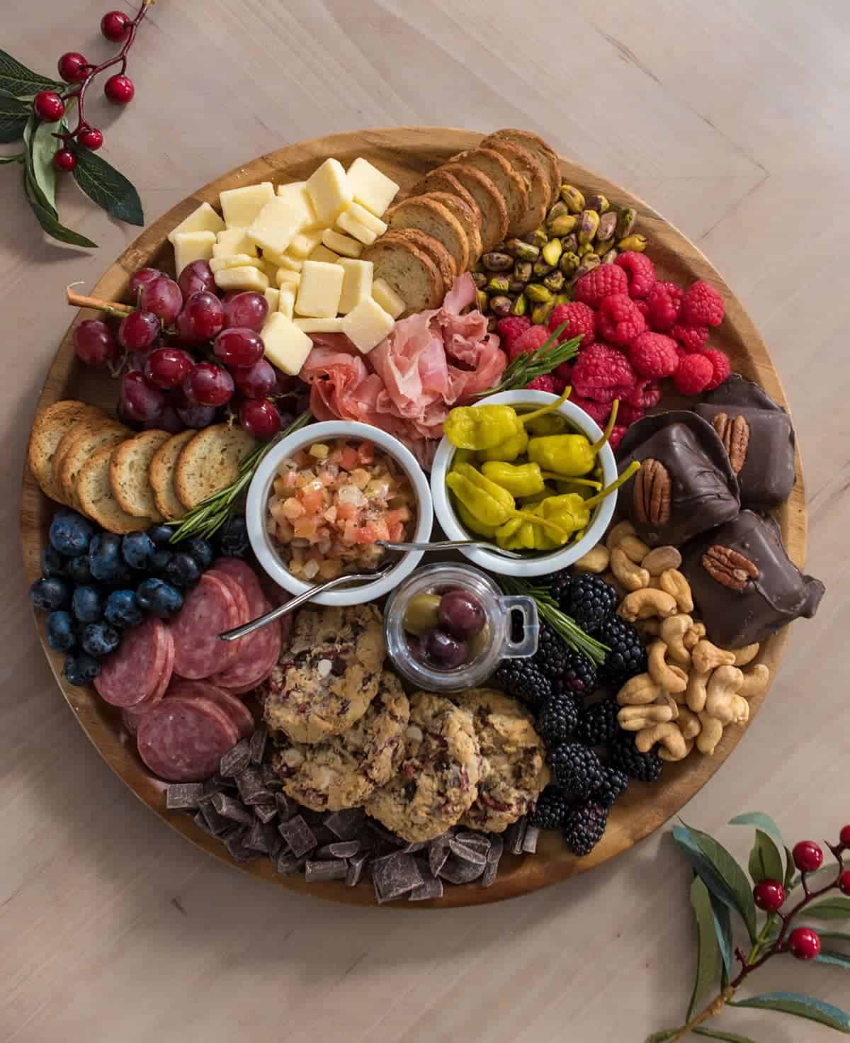 Charcuterie board on wooden platter featuring assortment of berries, cheese, nuts, bagel chips, chocolate, prosciutto, cranberry cookies, turtle brownies, bruschetta & pepporcinis.