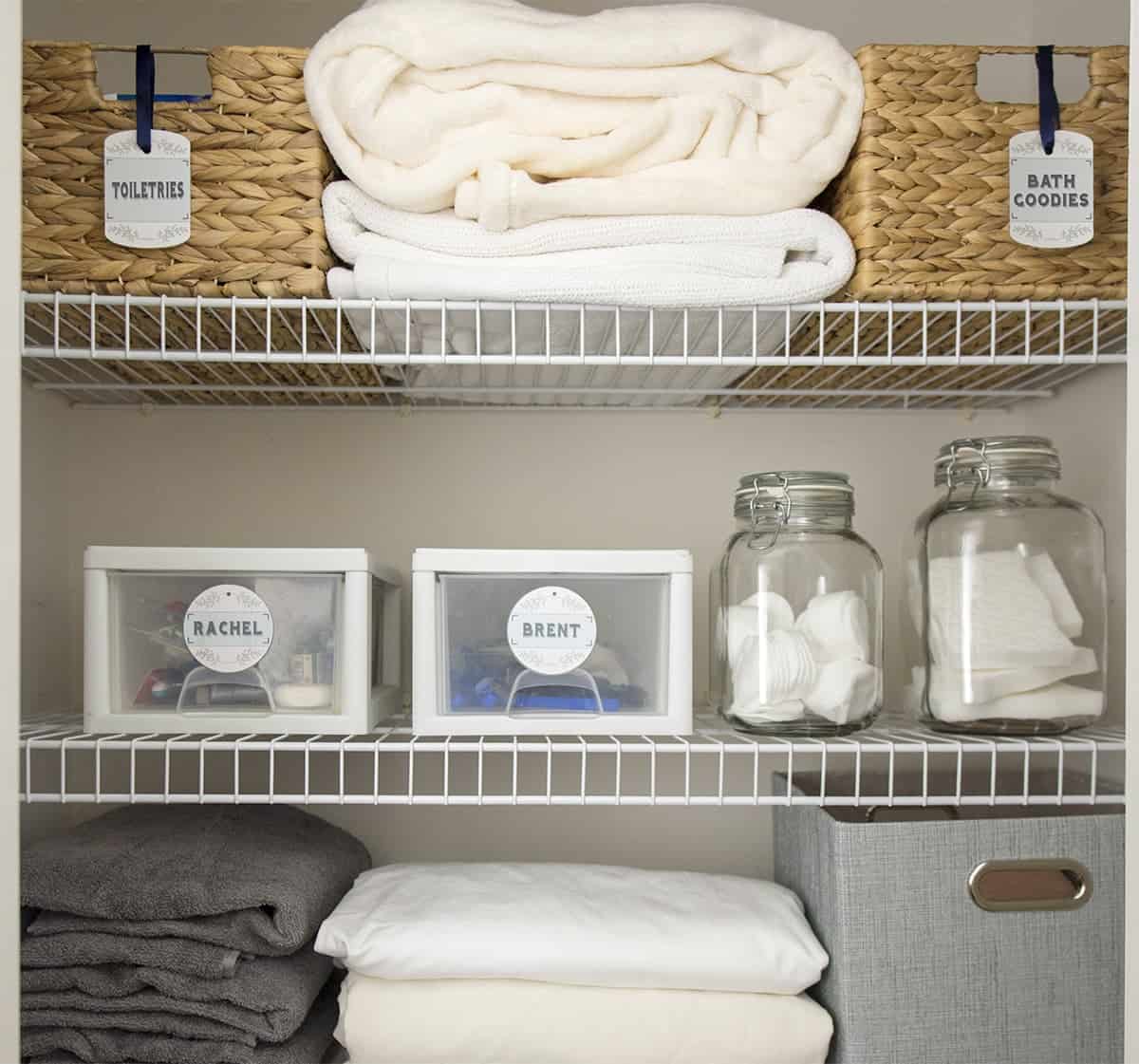 A closeup of an organized hallway linen closet with baskets and bins, towels, and organization labels.