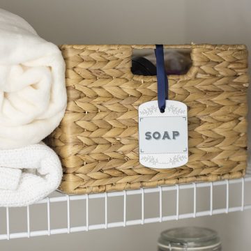 Printable label on wicker basket on white wire shelf next to white folded towels.