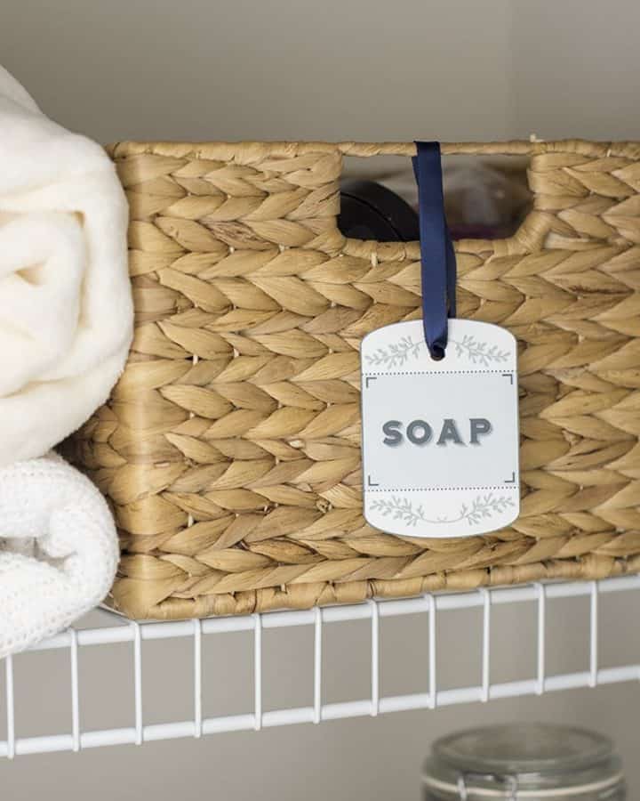 Printable label on wicker basket on white wire shelf next to white folded towels.
