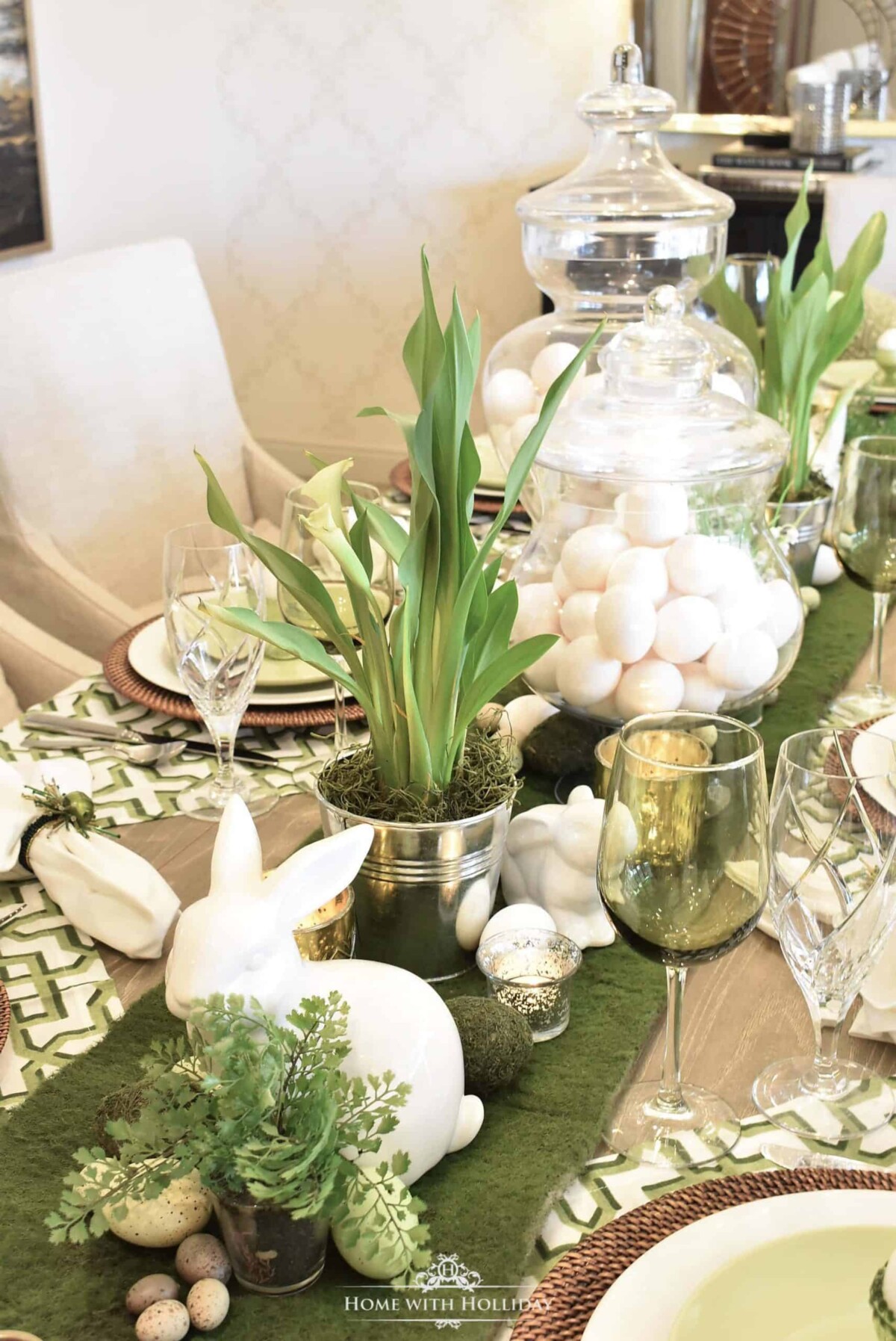 Elegant table setting with eggshell and green color scheme, green table runner, glass containers filled with eggs, and metal buckets with grass-like plants.