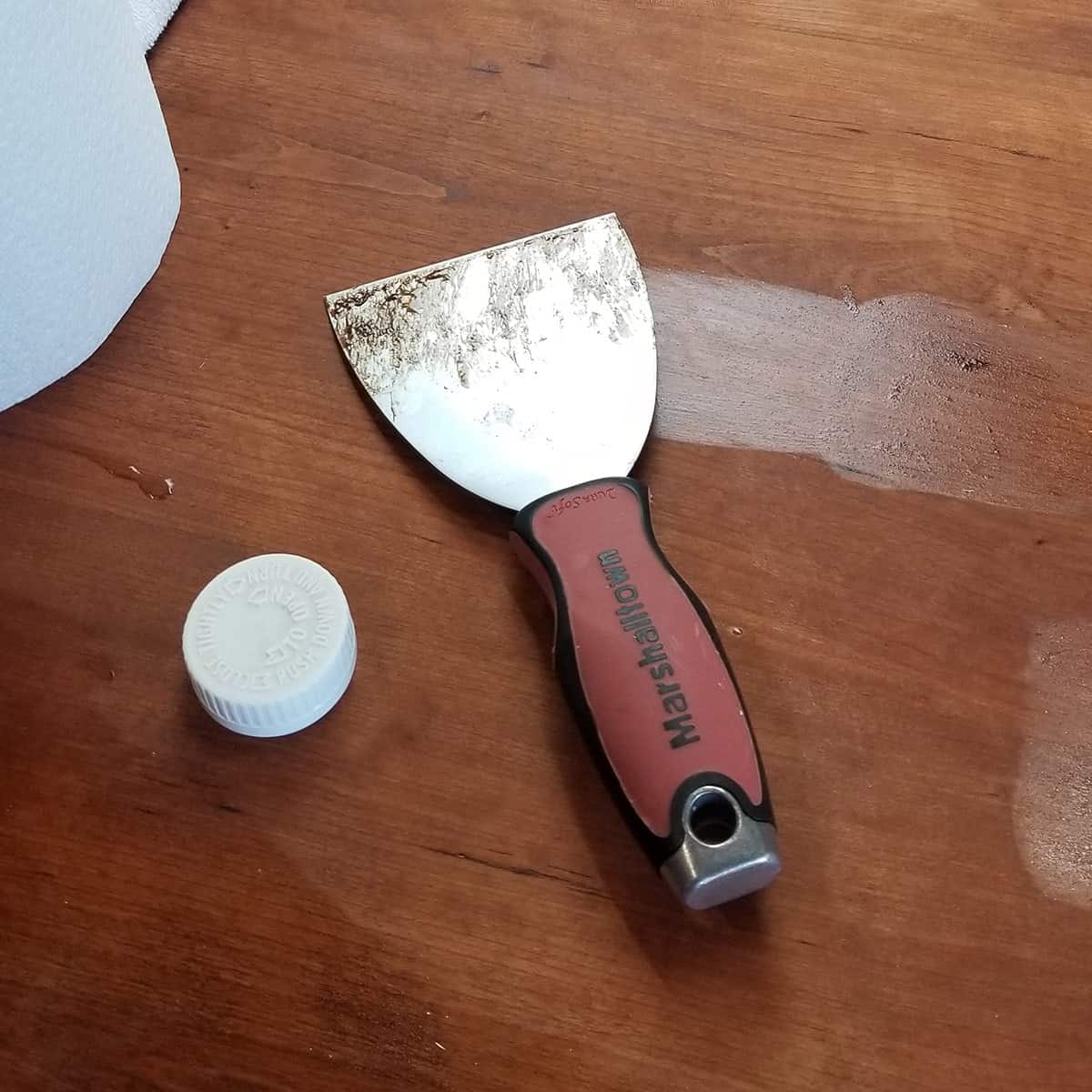 A paint scraper and furniture bleaching solution on cherry dining table.