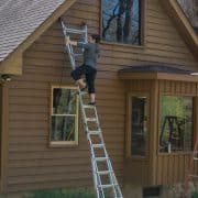 Woman on a ladder using a sprayer to stain a wood siding house exterior.
