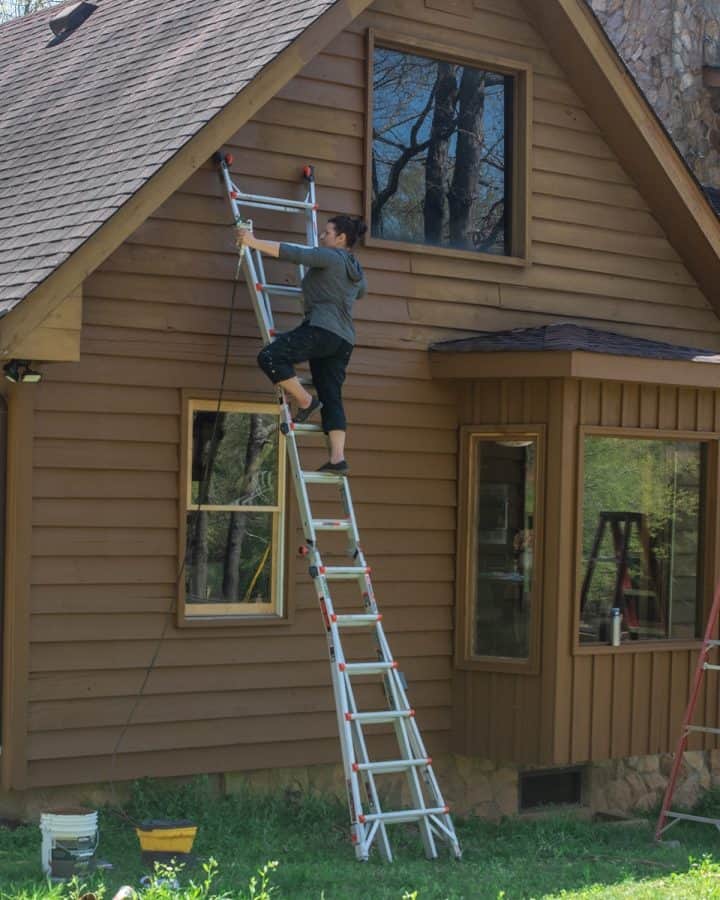 Woman on a ladder using a sprayer to stain a wood siding house exterior.