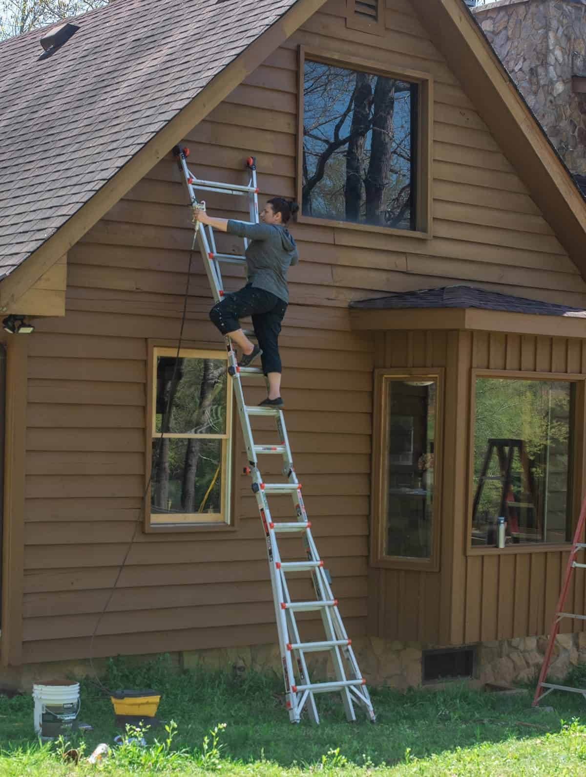 Side view of exterior of home midway through renovation. Woman on a ladder using paint sprayer to stain the home.