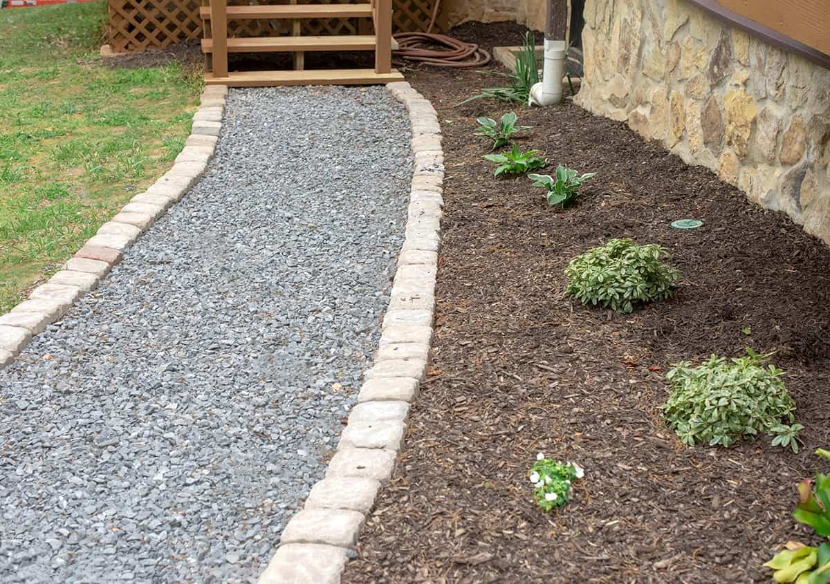 Paver stone border of loose gravel path walkway. Small garden with young plantings and fresh dark mulch sandwiched between house and path.