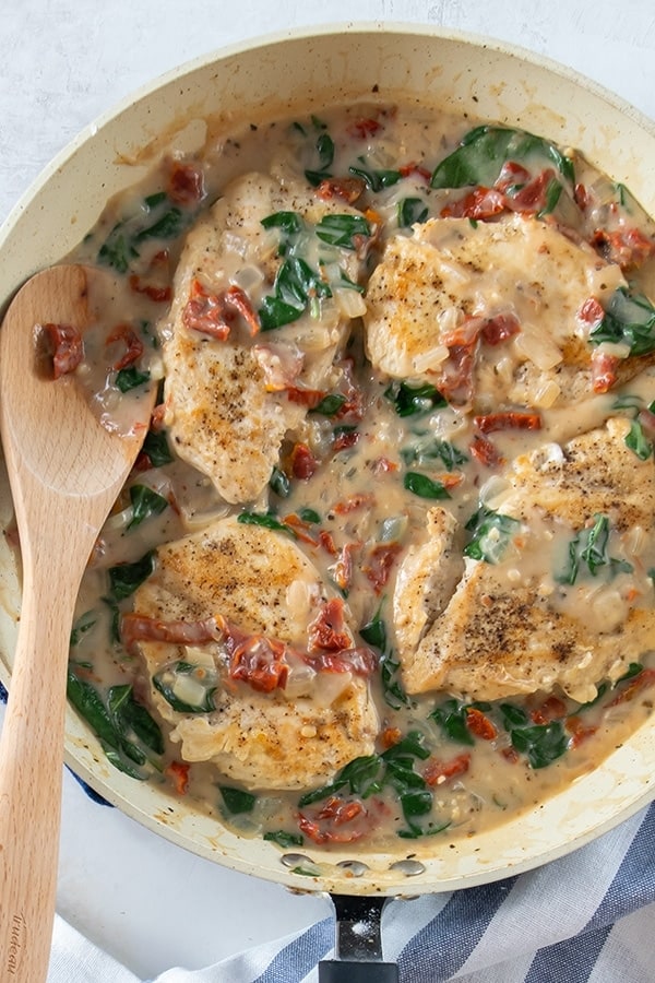 Keto Tuscan Chicken Recipe with Coconut Milk for creamy garlic sauce. Spinach, seasoned chicken and sun-dried tomatoes in the pan. Wooden spoon rests in pan.
