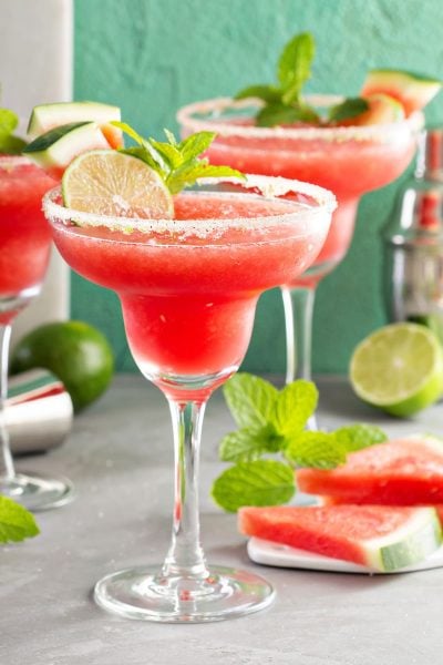 Frozen Watermelon margaritas with sugar rim, mint, limes and slices of watermelon in background.