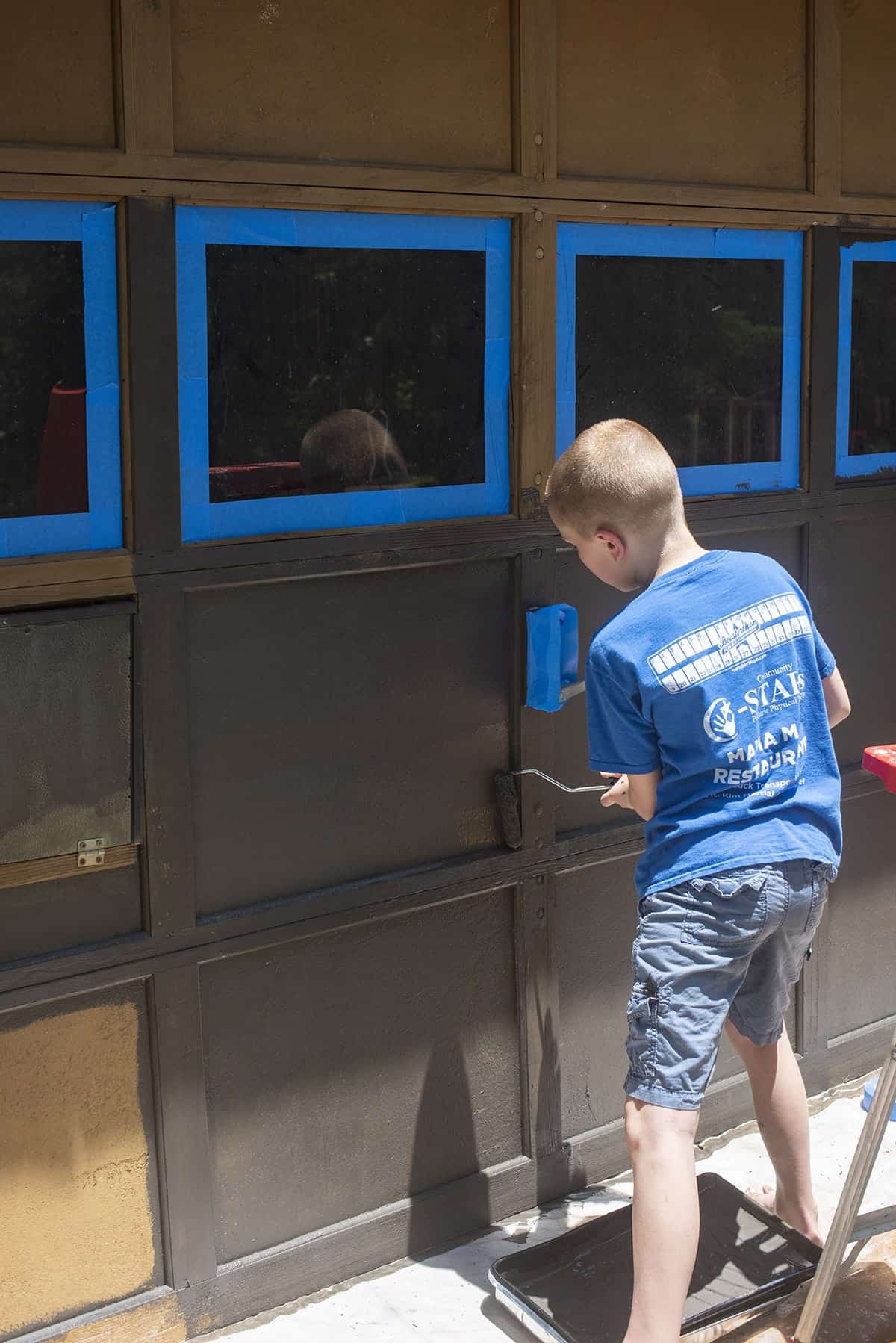 Taping around garage windows and boy painting a garage door with a roller.