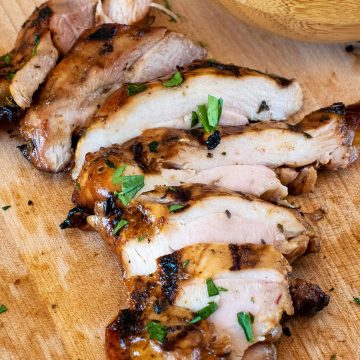 Juicy grilled and marinated chicken sliced on a wooden cutting board with a bowl of fresh herbs in the background.