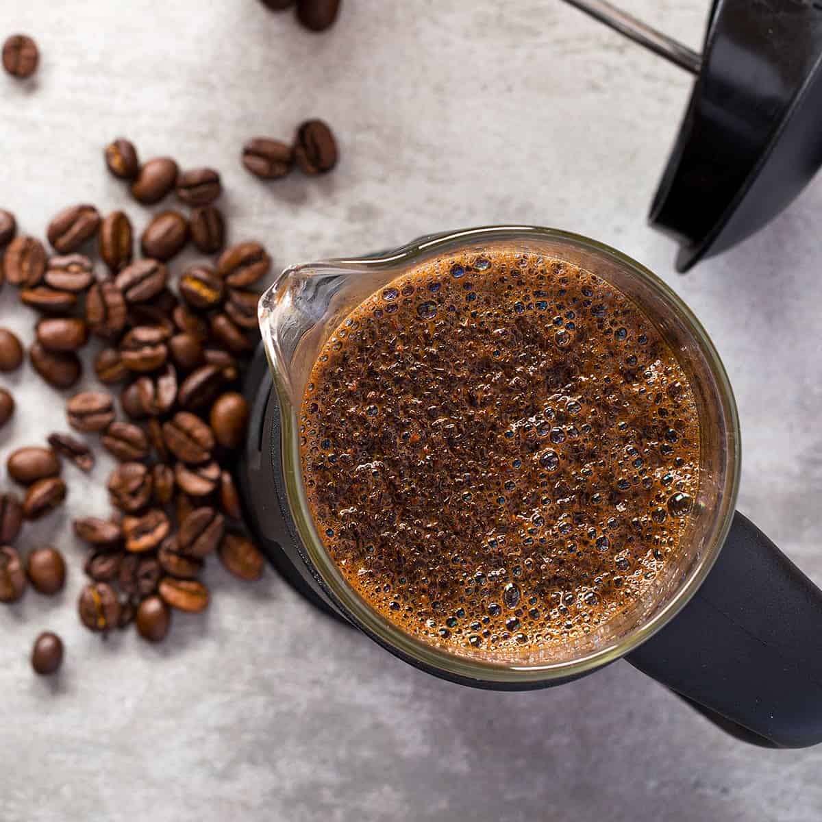 A look down at coffee as it is brewing. The grounds are wet with coffee beans sprinkled on a grey background.