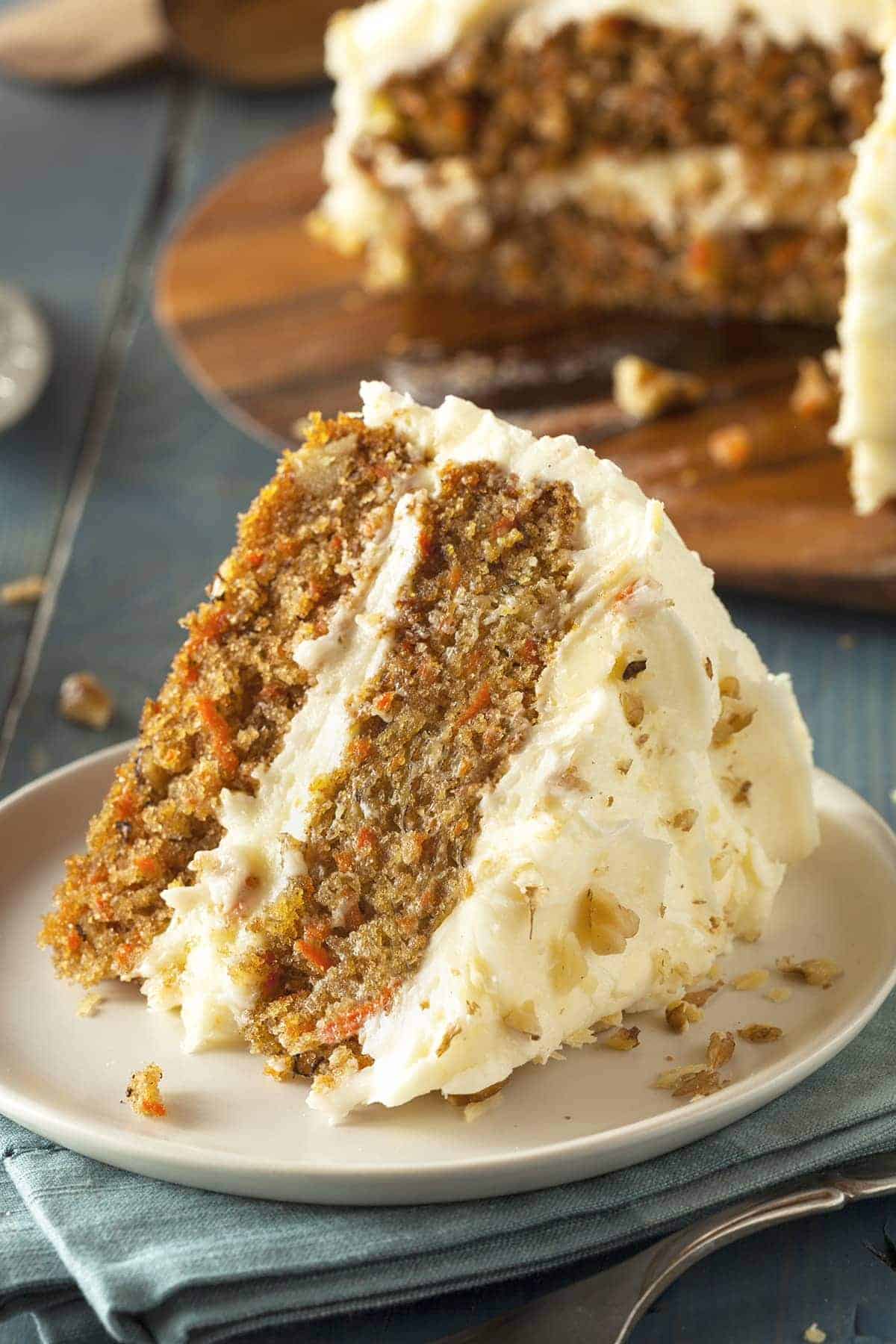 Slice of carrot cake covered in cream cheese frosting, garnished with chopped walnuts on white plate over blue towel and fork. 