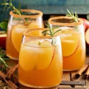 Side view of three glasses of caramel apple sangria on a rustic wood board. Cinnamon sticks lay around the glasses with slices of apple and herbs as garnish.