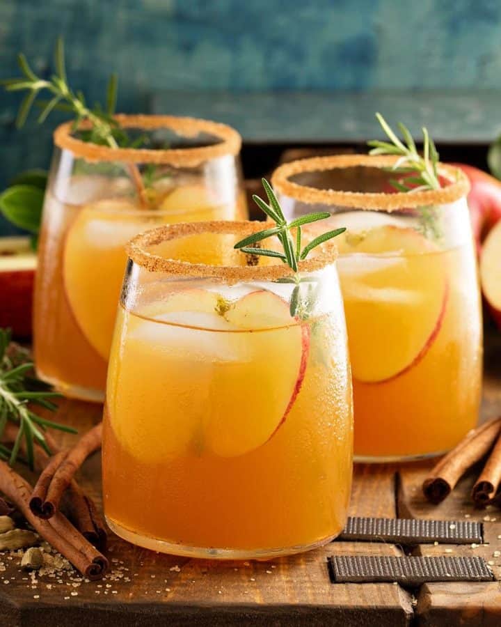 Side view of three glasses of caramel apple sangria on a rustic wood board. Cinnamon sticks lay around the glasses with slices of apple and herbs as garnish.