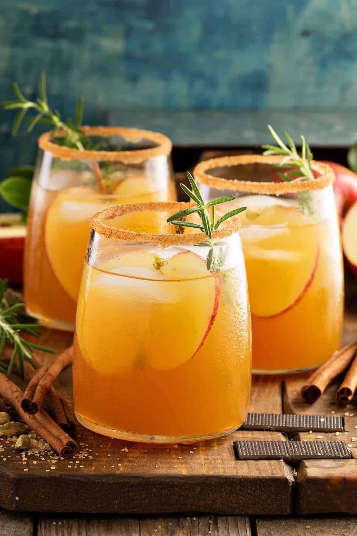 Side view of three glasses of caramel apple sangria on a rustic wood board. Cinnamon sticks lay around the glasses with slices of apple and herbs as garnish.