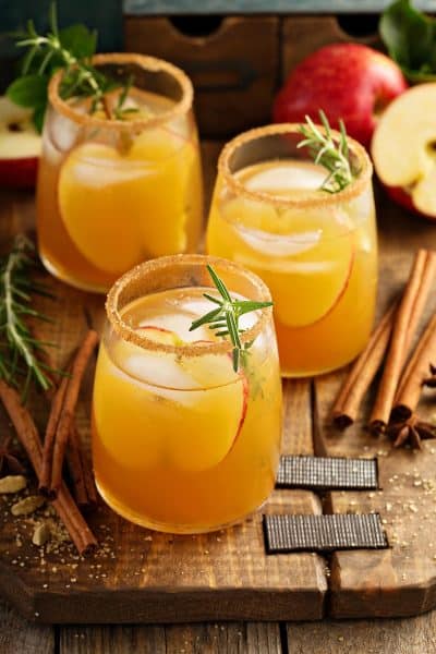 Three caramel apple sangria in glasses on a wood board. The glasses are garnished with an apple slice, rosemary, and a sugar rim.