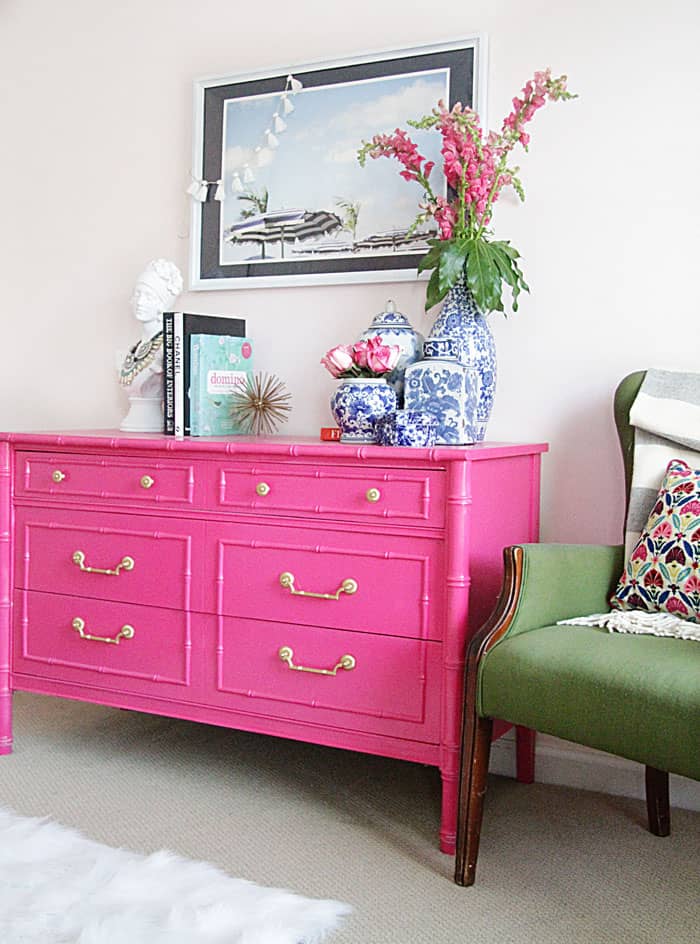Bright pink dresser with gold drawer pulls next to green settee with patterned pillow and gray throw.