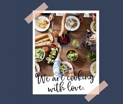 Polaroid of people gathered around a table set with food and passing the food around. Text Overlay reads we are cooking with love.