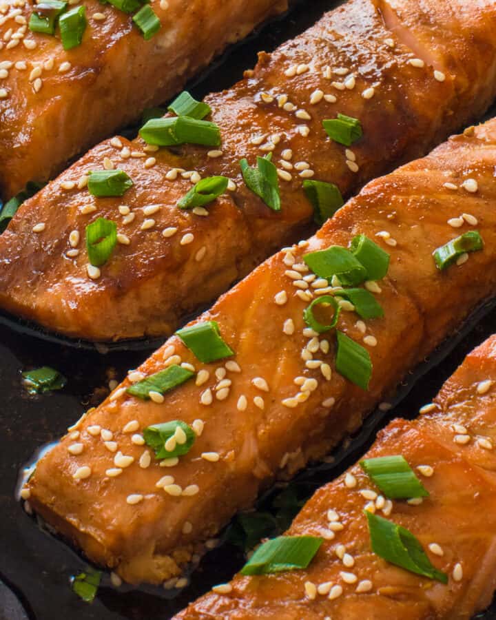 Close up of four pieces baked Salmon in a pan in Teriyaki sauce with sesame seeds and chives garnish.