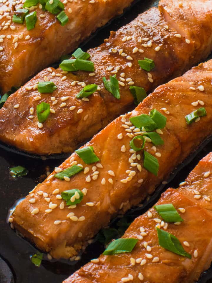 Close up of four pieces baked Salmon in a pan in Teriyaki sauce with sesame seeds and chives garnish.