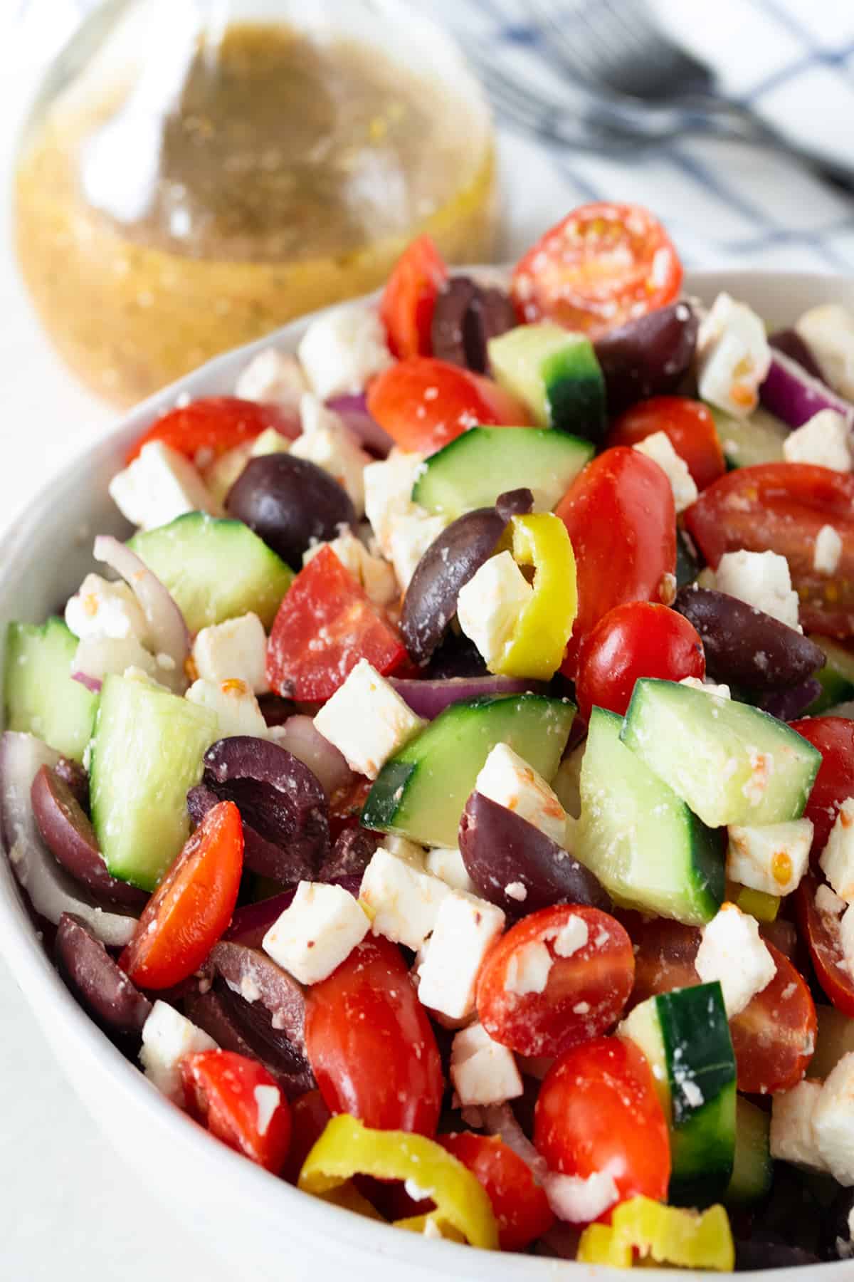 Closeup of chopped greek salad with olives, tomatoes, cucumber, and feta with dressing in the background.