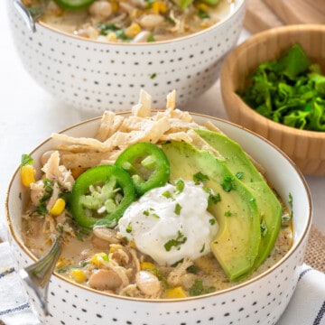 Two Bowls of white chicken chili on white background. Chili is garnished with tortilla strips, jalapeno, avocado, and cilantro.