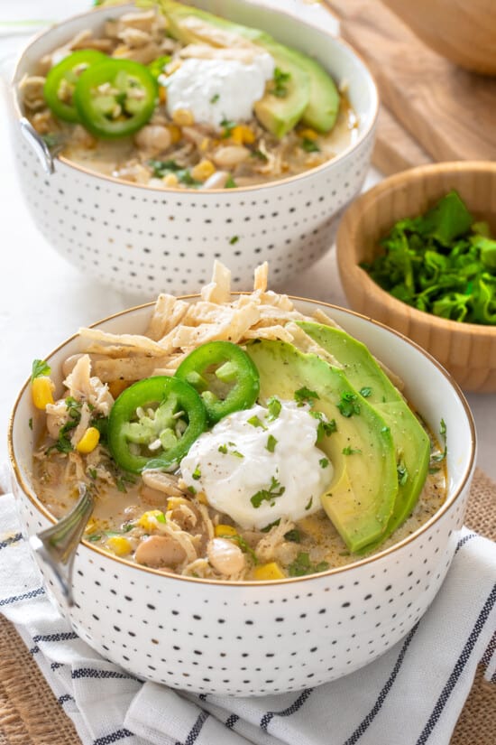 Two Bowls of white chicken chili on white background. Chili is garnished with tortilla strips, jalapeno, avocado, and cilantro.