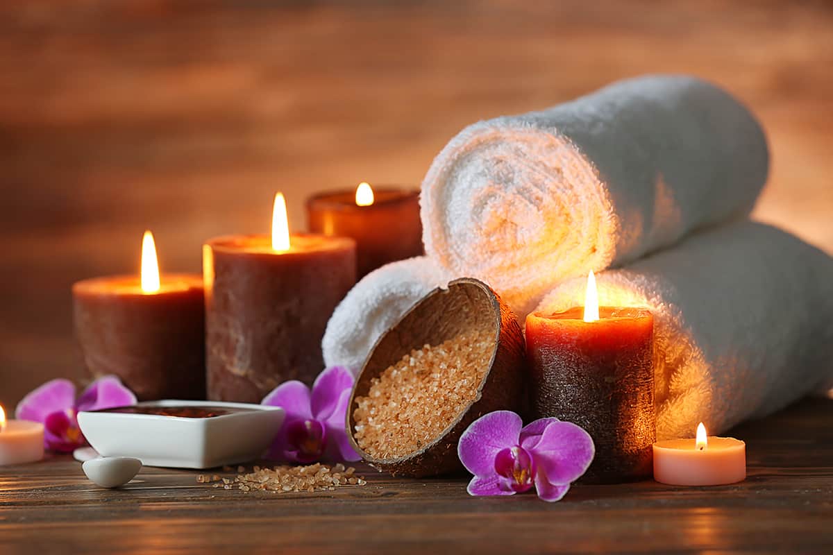 A grouping of spa products including lit candles, folded towels, salt scrub, and orchids on a wood background.