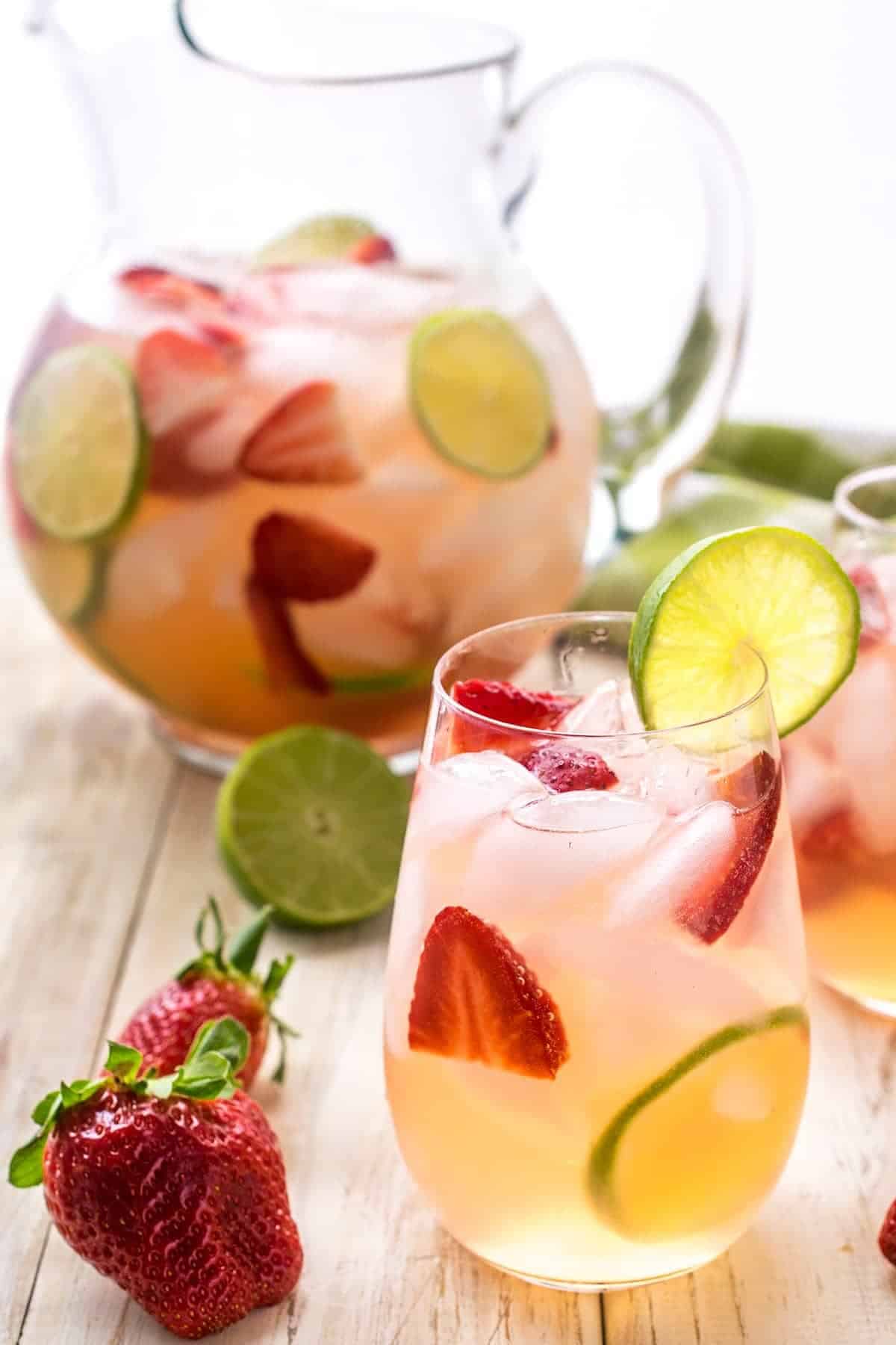 Strawberry sangria in a glass with lime garnish. Pitcher of strawberry sangria in the background. Whole strawberry and half lime on table top.