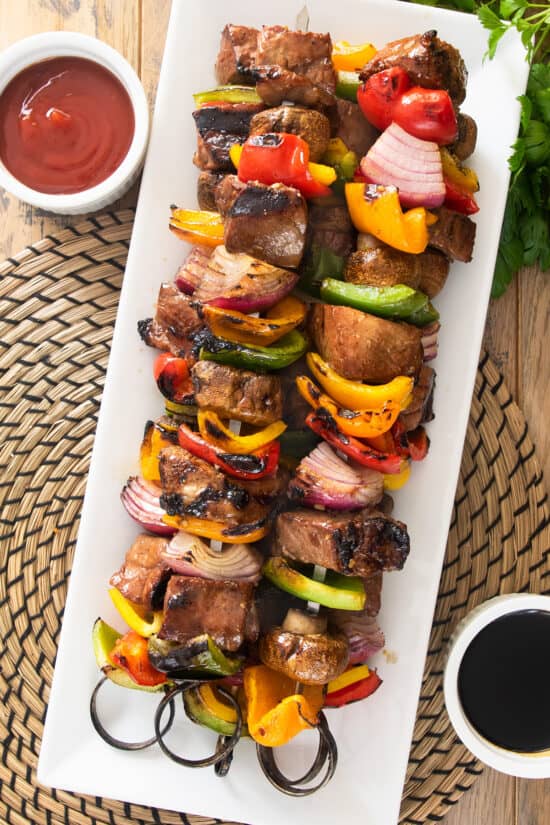 Skewered steak kabobs with peppers, mushrooms, and onions on a long serving platter.