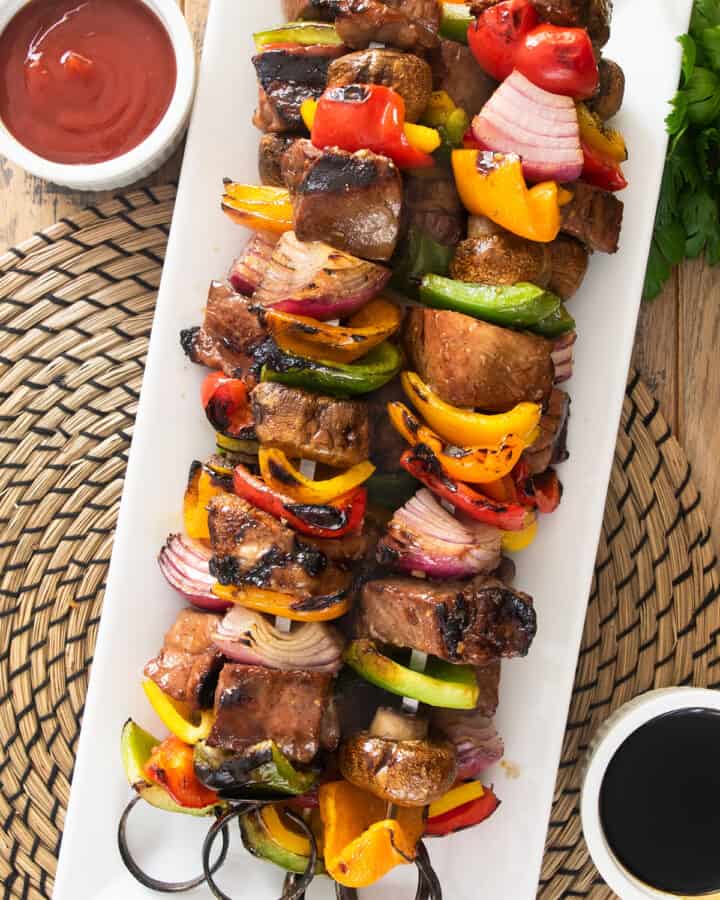 Skewered steak kabobs with peppers, mushrooms, and onions on a long serving platter.
