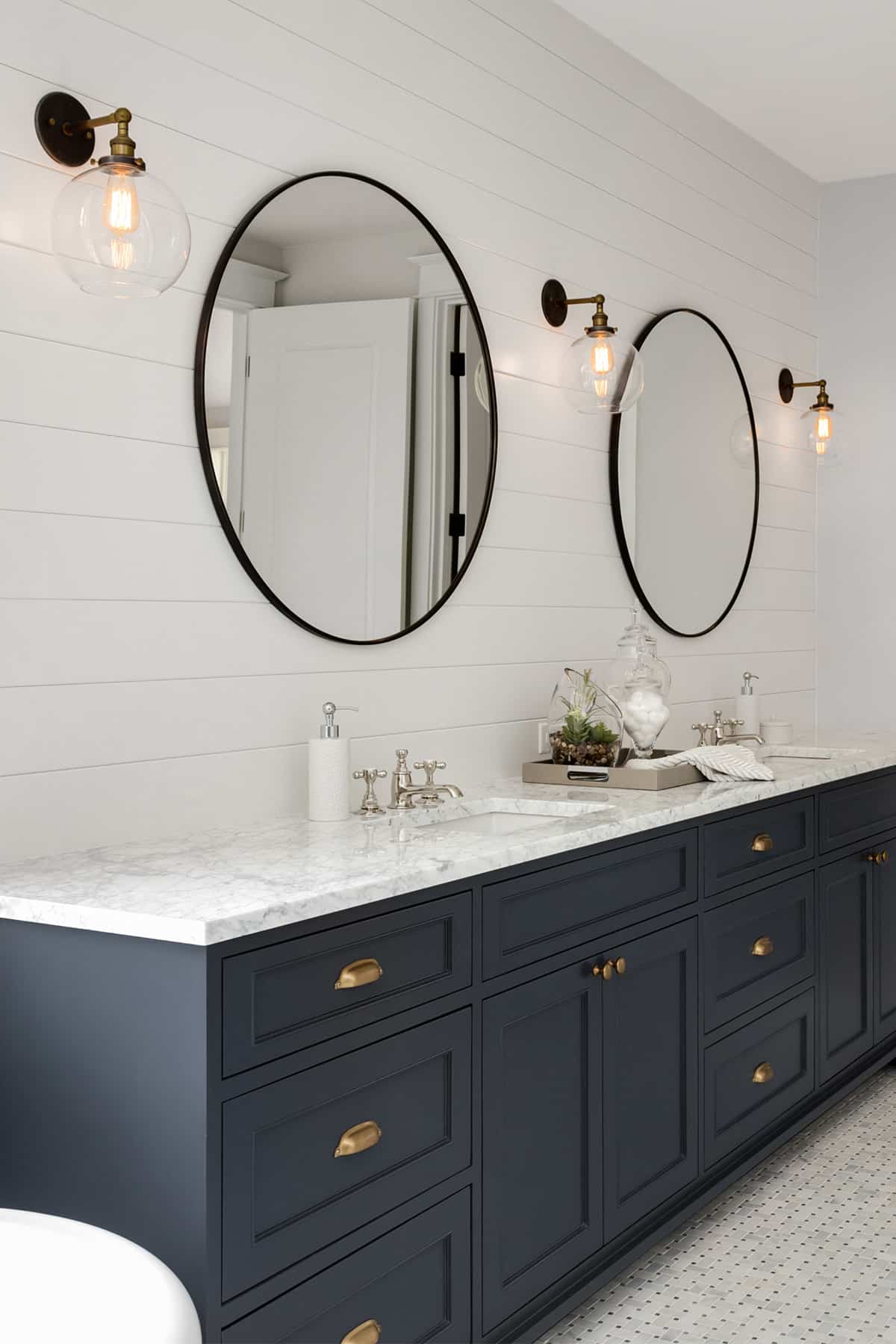 White bathroom with navy vanity, shiplap, and brass sconces to the side of round mirrors.
