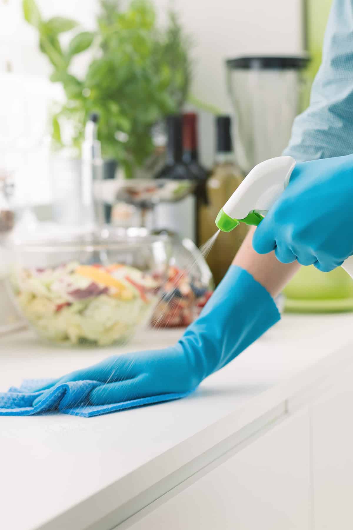Woman cleaning countertop with gloves on using a spray bottle and sponge.