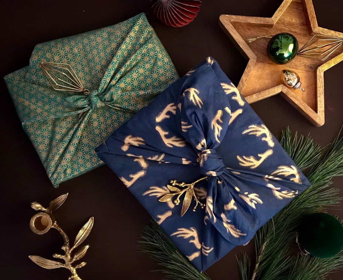 Fabric wrapped gift packages with ornaments on them and greenery scattered on a table.