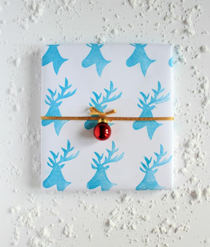Christmas gift wrapped in white paper with blue reindeer stamps and a gold ribbon with red bell.