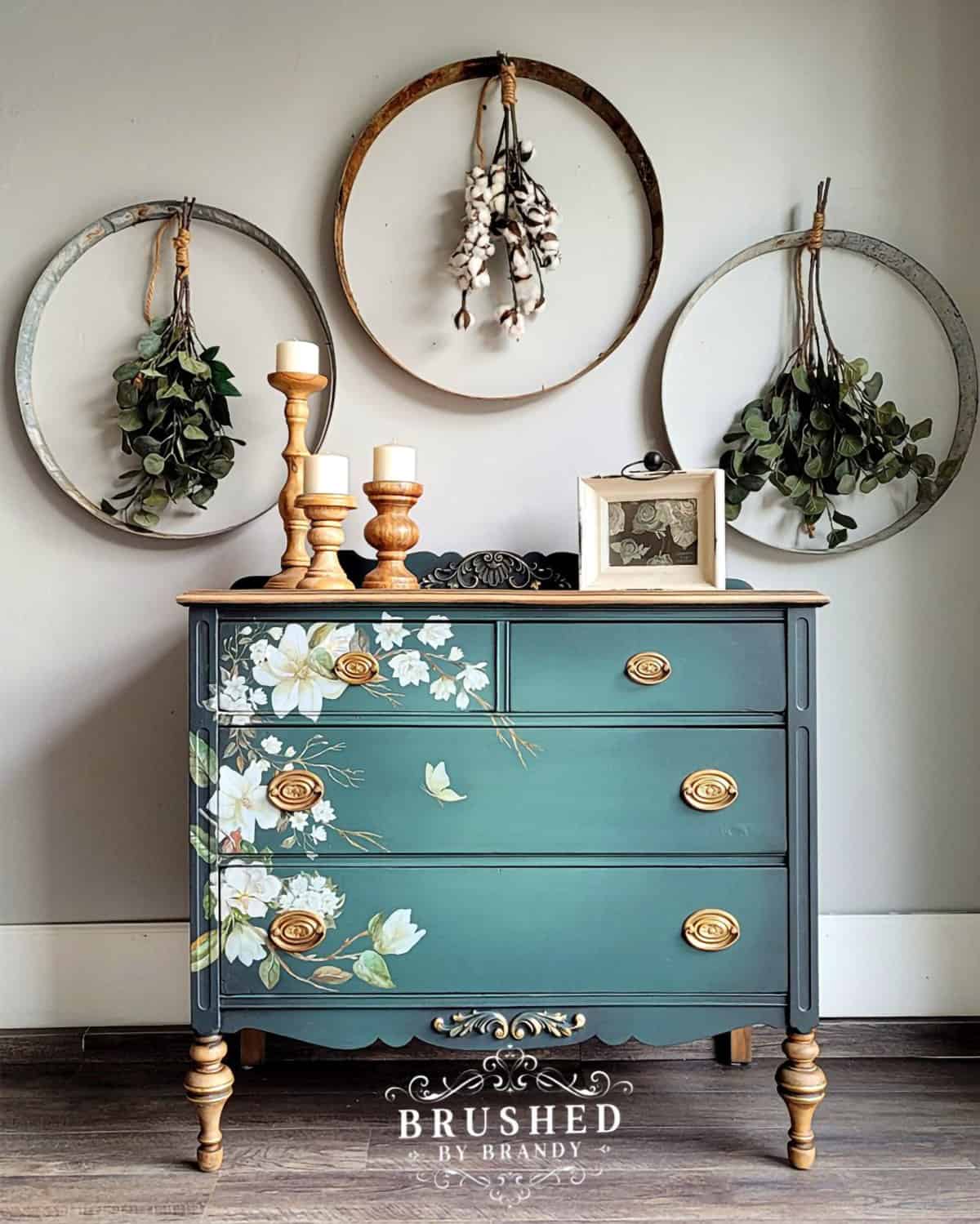 Juniper green dresser with brass hardware and floral design. Large hoops with dried herbs hang on wall in background.