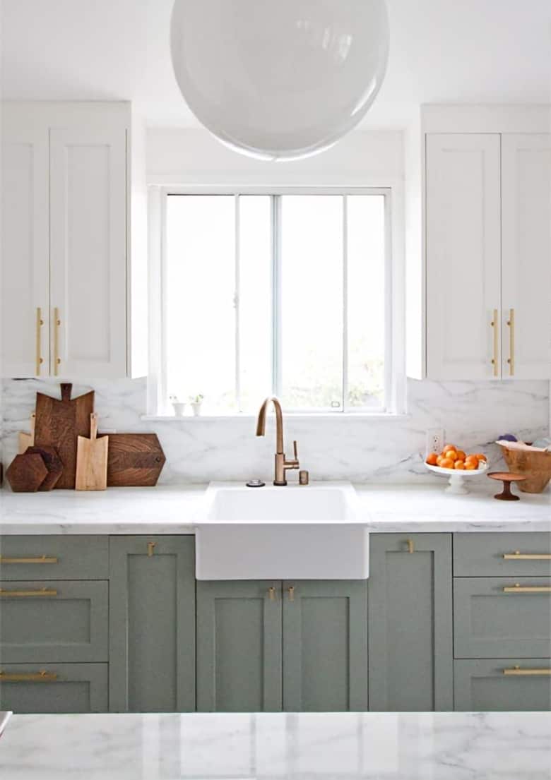 Modern kitchen with dual white and muted green cabinetry, brass hardware, marble backsplash, and large white sphere light. 