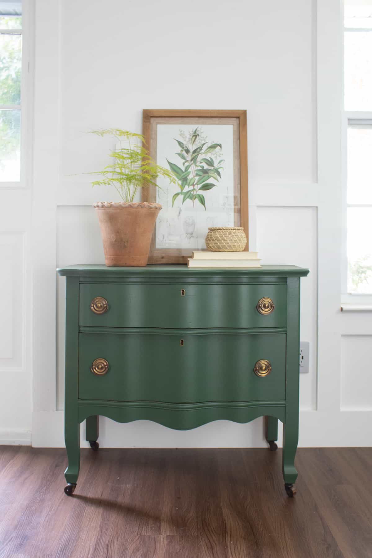 Vine green antique dresser with brass pulls and legs against white wood background wall with decorated top.
