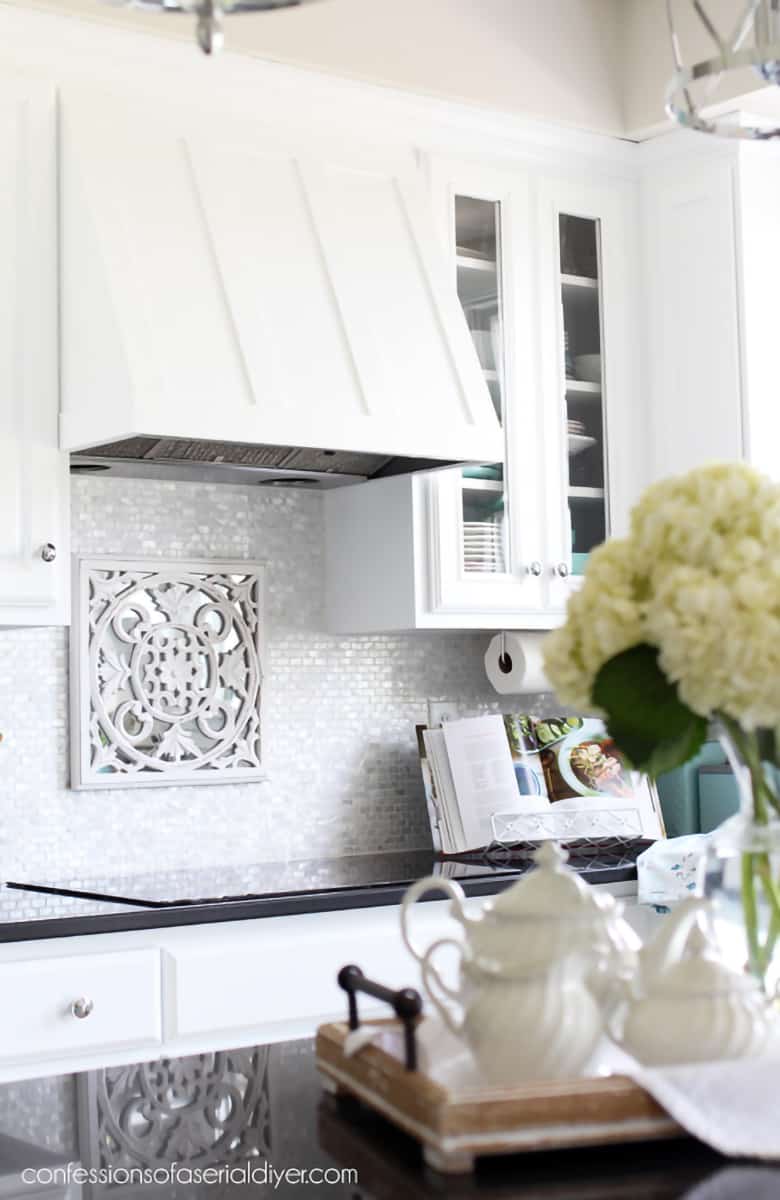 DIY Range Hood Cover painted to blend in seamlessly with white cabinets in French country style kitchen featuring light reflective tiled backsplash and black countertops.