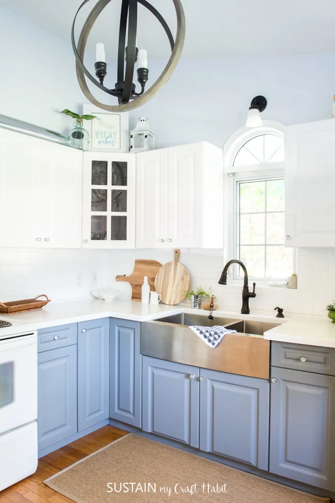 A Kitchen featuring blue cabinets, brushed metal apron style double sink with window view and white countertops and backsplash.