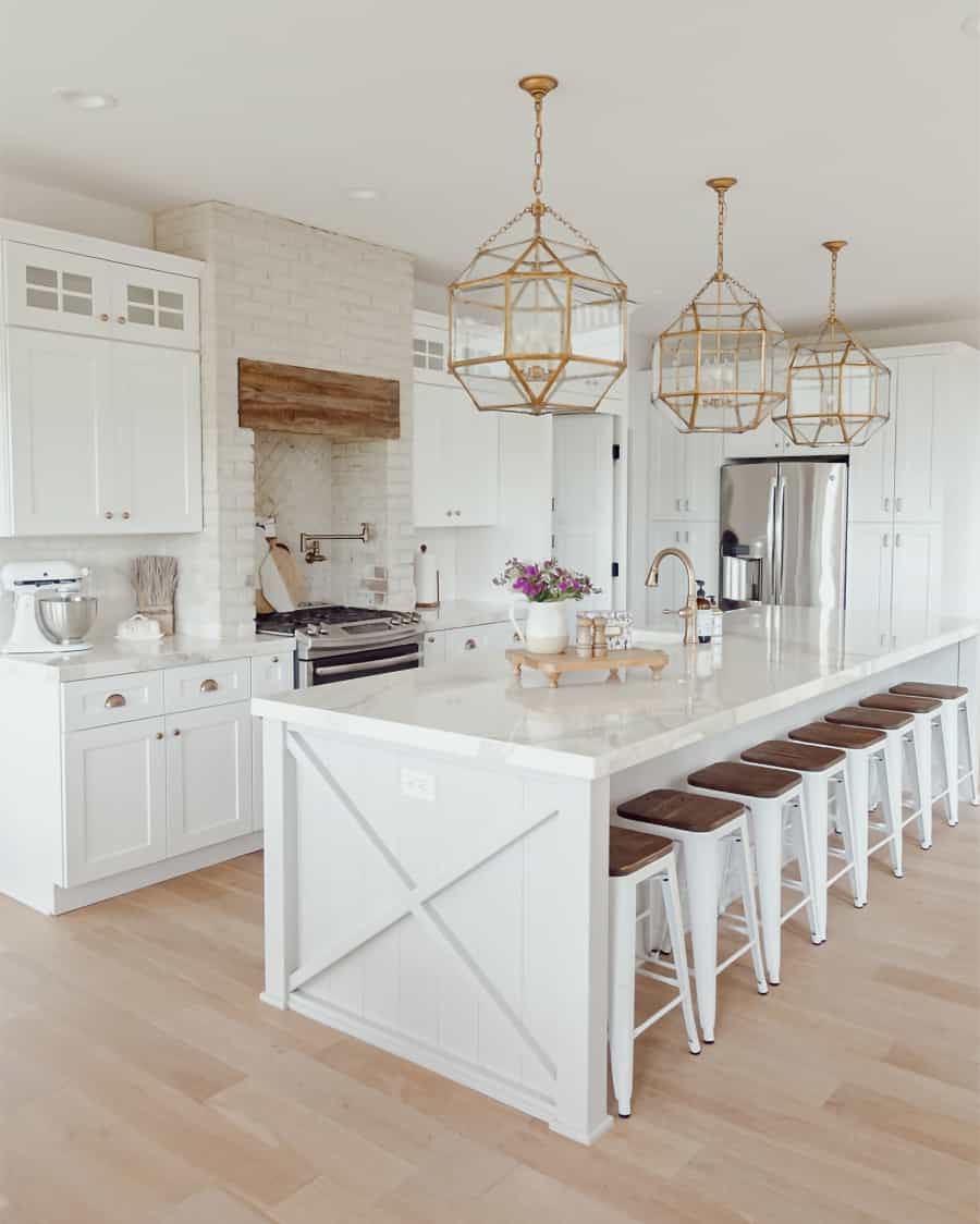 Elegant and spacious white kitchen with oversized marble island, brass pendant lights, and wooden hood and barstool seats. 