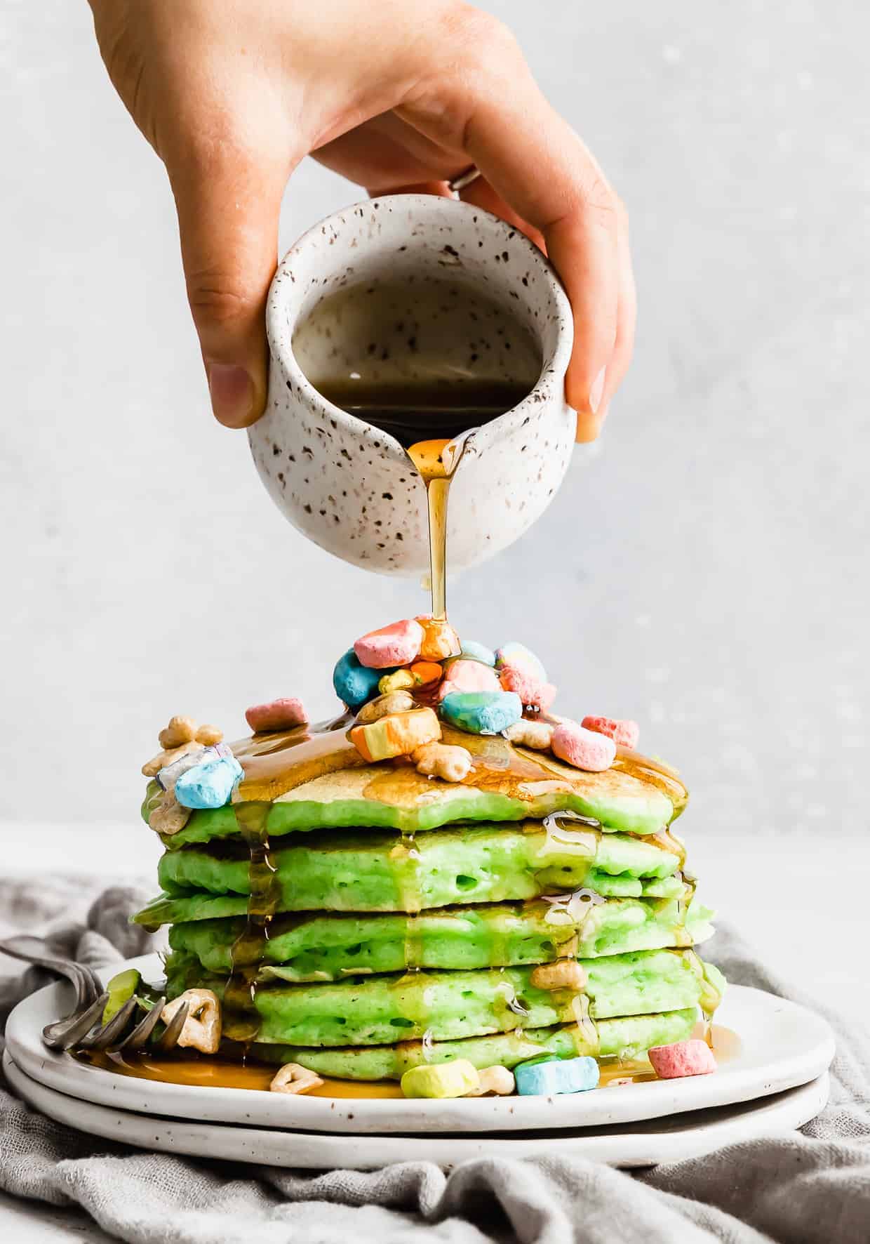Hand pouring syrup over green pancake stack with lucky charms marshmallows over top.