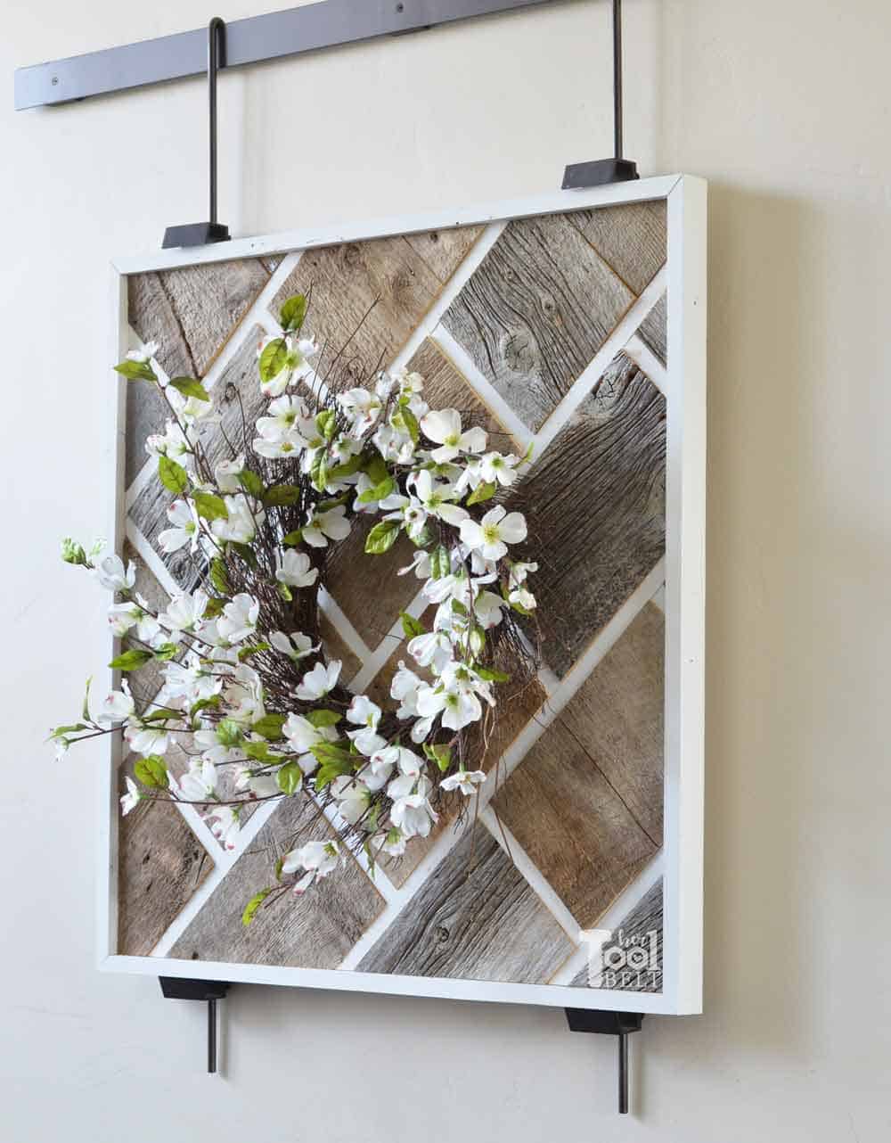 Reclaimed barnwood planks framed wall art suspended from a black metal rod on a beige wall with a flower wreath.