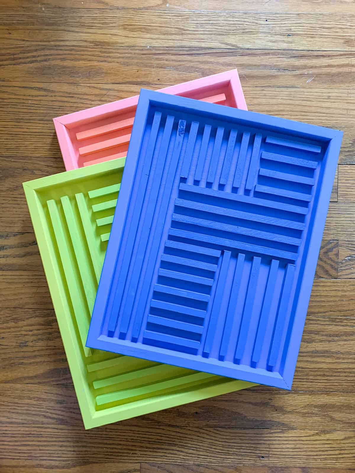 A bright pink, green, and blue wood wall art pieces with geometric patterns stacked against a wood grain table backdrop.