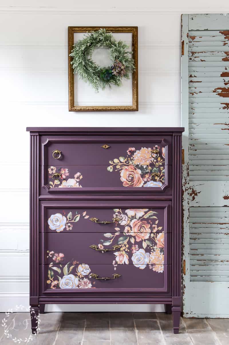 A painted purple dresser with delicate floral design in front of white plank wall with simple wreath. 