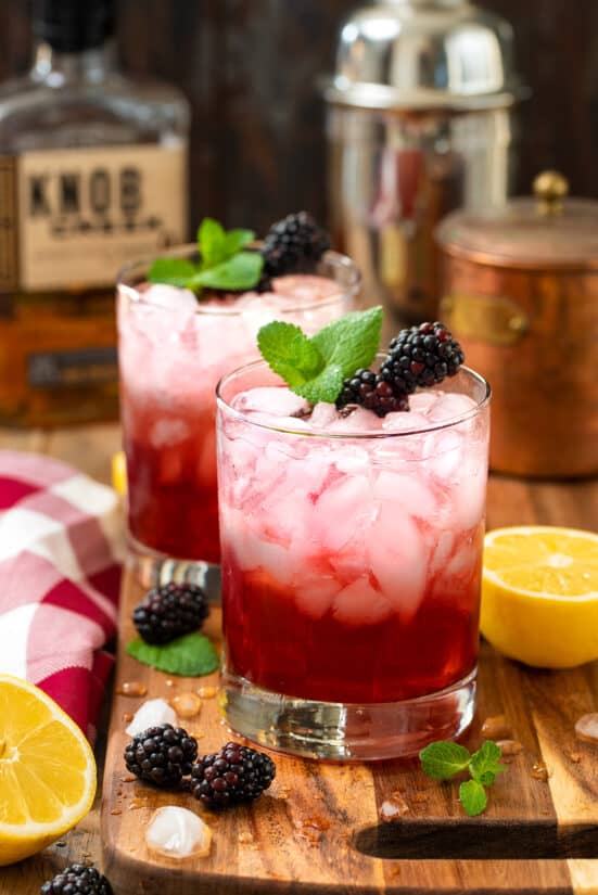Two glasses of blackberry bourbon smash cocktail garnished with mint and blackberries on a wood table.