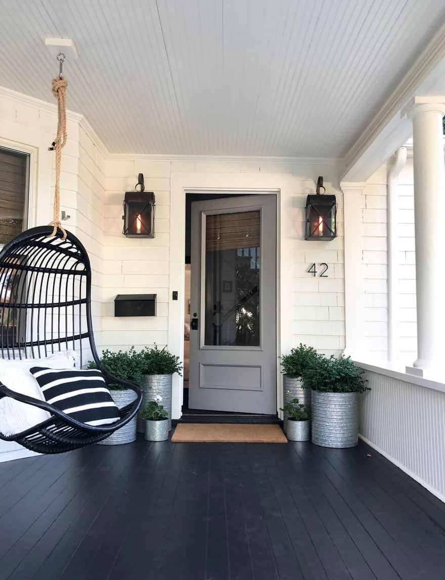 Single seat hanging black rattan porch swing with black and white pillows on black stained front porch