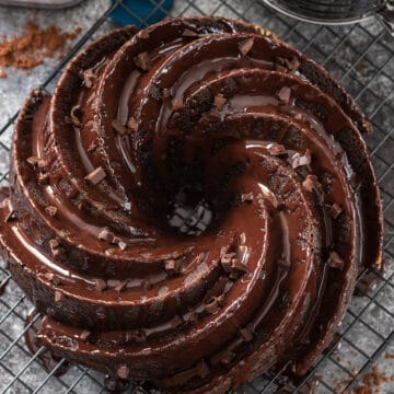 Whole chocolate bundt cake with chocolate ganache drizzle and flakes on a table to cool.