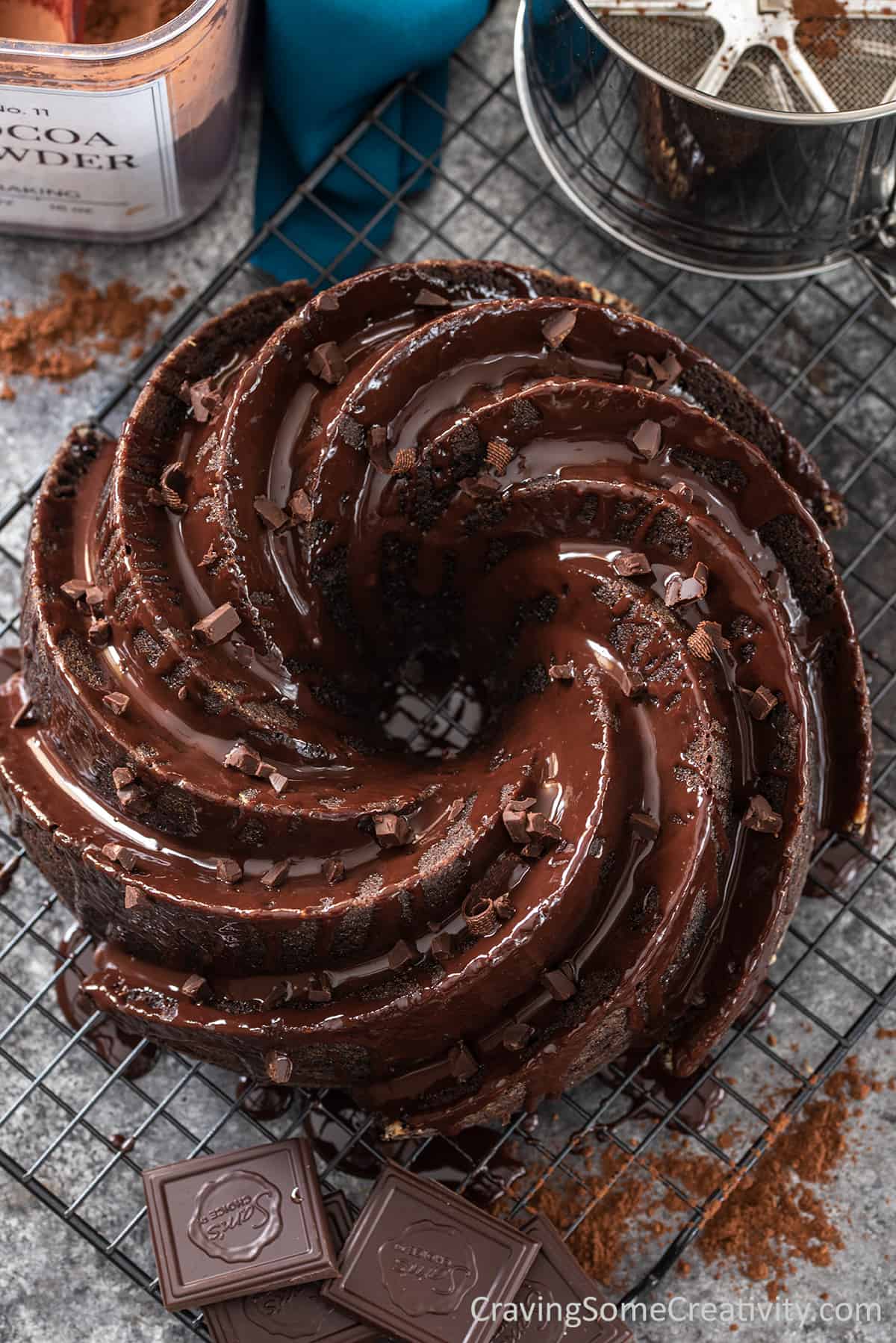 Whole chocolate bundt cake with chocolate ganache drizzle and flakes on a table to cool.