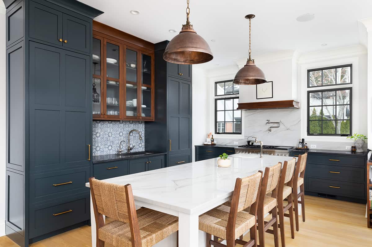 Behr Midnight blue painted cabinets in a classic old world kitchen with modern white accents and backsplash. Wicker chairs, copper light fixtures, and wood floors add warmth.